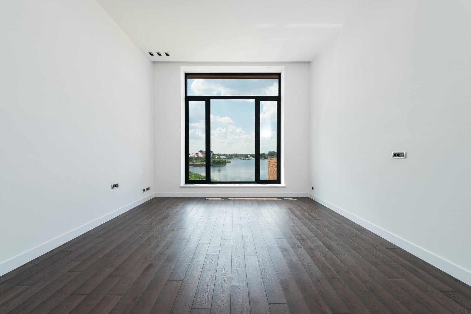 An empty residential room with a hardwood floor and a tall window overlooking a lake